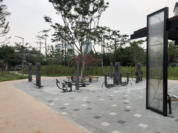 Outdoor-Park mit verschiedenen Fitnessgeräten, Pflanzen und urbanen Elementen unter einem Himmel-Hintergrund.
