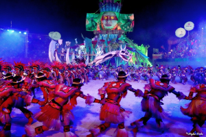 Eine Gruppe von Menschen in farbenfrohen Kostümen tanzt auf einer Bühne vor einem sitzenden Publikum, mit Lichtern, Pfählen, Ballons und einem Bildschirm, der eine Person in den Hintergrund zeigt, unter einem sichtbaren Himmel, bei der Rio-Karneval 2014.