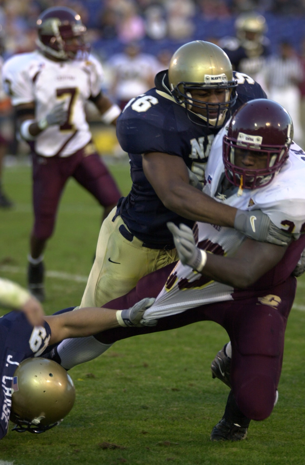 Zwei Fußballspieler in Helmen und Uniformen tackeln sich gegenseitig auf dem Feld, während andere zusehen.