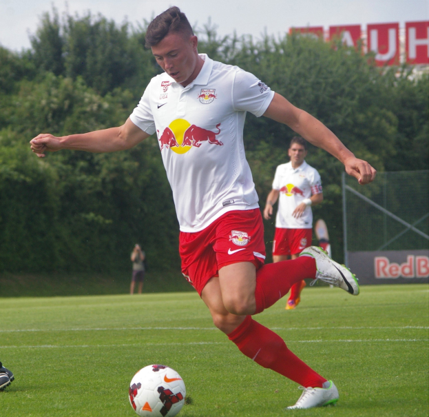 Ein professioneller Fußballspieler schießt einen Ball auf einem grünen Feld mit "RB Leipzig" im Hintergrund unter einem klaren blauen Himmel.