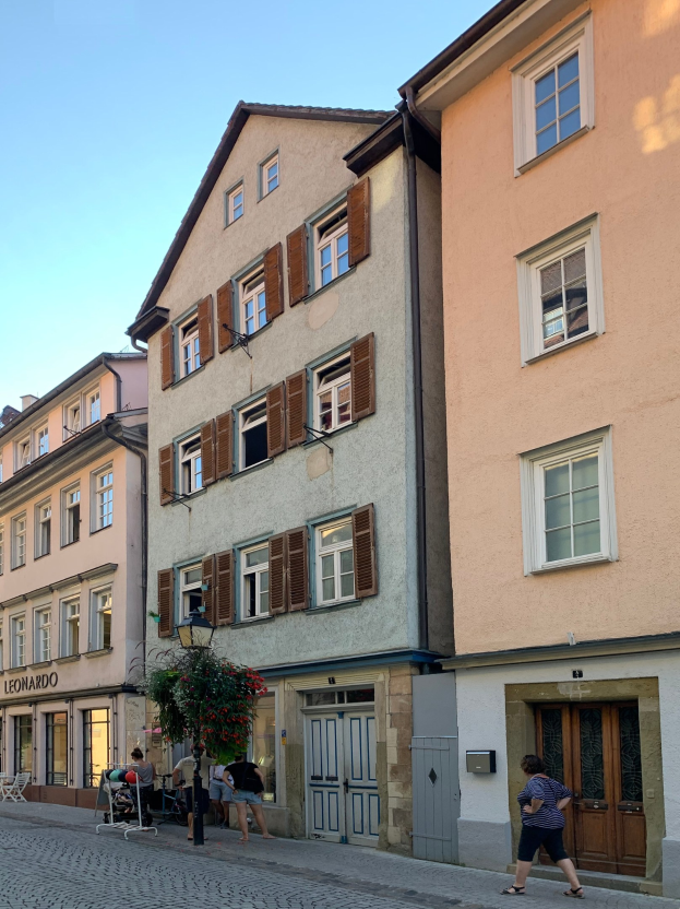 Eine Frau spaziert auf einem Kopfsteinpflasterweg in der Altstadt von Heidelberg, umgeben von hohen Gebäuden, einer Straßenlaterne mit einer Pflanze, verstreuten Stühlen und Tischen und dem Himmel im Hintergrund.