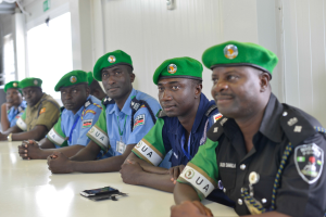 Eine Gruppe von Polizeibeamten sitzt an einem Tisch mit Mobiltelefonen und anderen Gegenständen in einem Raum mit einer Wand, Fenstern links und einer beleuchteten Decke.