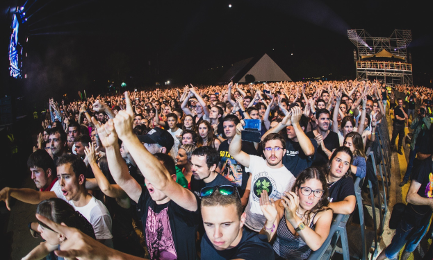 Menschenmenge auf einem Musikfestival mit erhobenen Händen, sitzend und stehend, Metallzaun auf der rechten Seite, Gebäude und Lichter im Hintergrund unter einem dunklen Himmel.