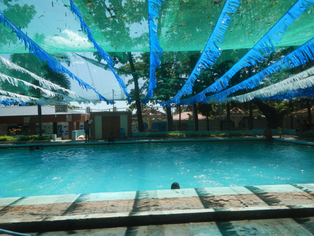 Ein großes Schwimmbecken mit blauen und grünen Bändern, die von der Decke hängen, umgeben von Häusern, Bäumen und einem klaren blauen Himmel, mit Menschen, die im Wasser und in der Sonne genießen.