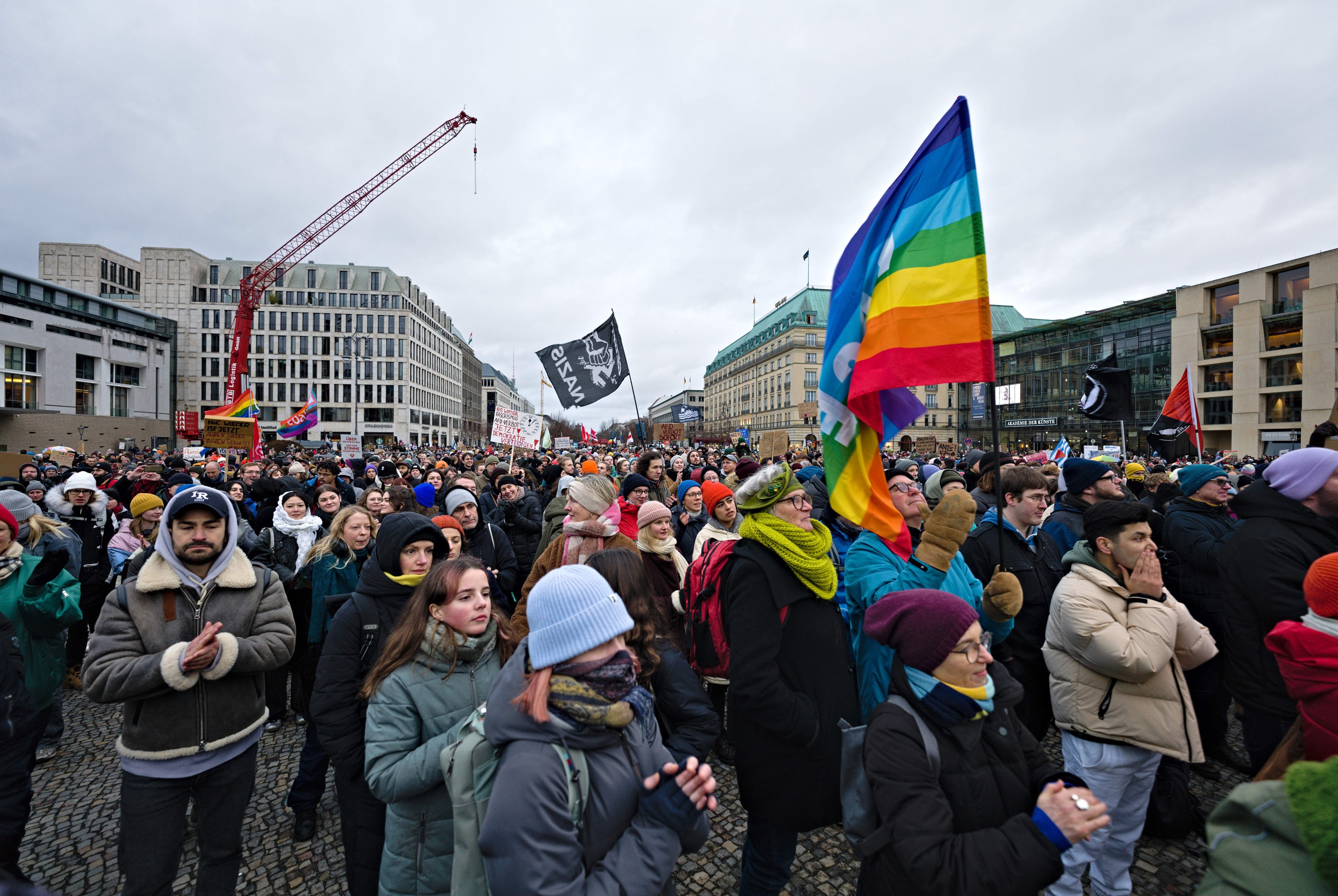 Eine große Gruppe von Menschen steht vor einem Gebäude, hält Fahnen und Transparente mit der Aufschrift "Lgbtq+ Rechte Demonstration in Berlin", einige tragen Mützen und Taschen, im Hintergrund sind Gebäude mit Fenstern, ein Kran und ein bewölkter Himmel zu sehen.