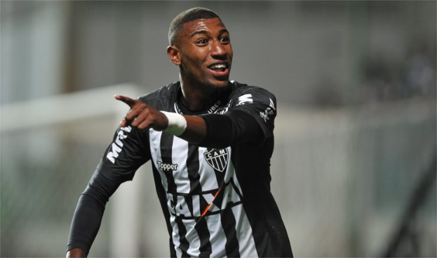 Ein Fußballspieler in einem schwarz-weißen gestreiften Trikot feiert mit einem Lächeln nach einem Tor, der Hintergrund ist unscharf, um den Moment zu betonen.