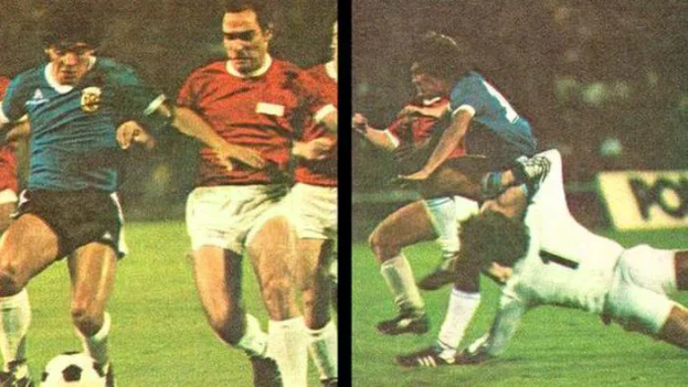 Eine Gruppe von Männern, die Fußball auf einem Feld spielen, wobei einer den Ball tritt und andere versuchen, die Kontrolle zu erlangen, vor einer Tafel in einem Stadion.