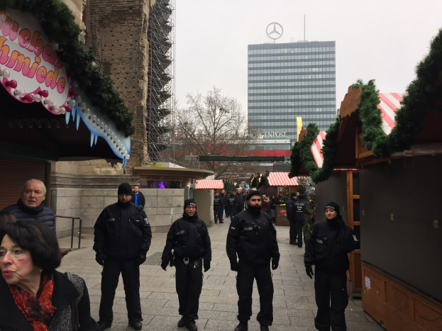 Polizeibeamte in schwarzen Uniformen stehen vor einem geschmützten Weihnachtsmarkt in Berlin, mit festlichen Dekoelementen, Gebäuden, Bäumen und einem klaren blauen Himmel im Hintergrund.