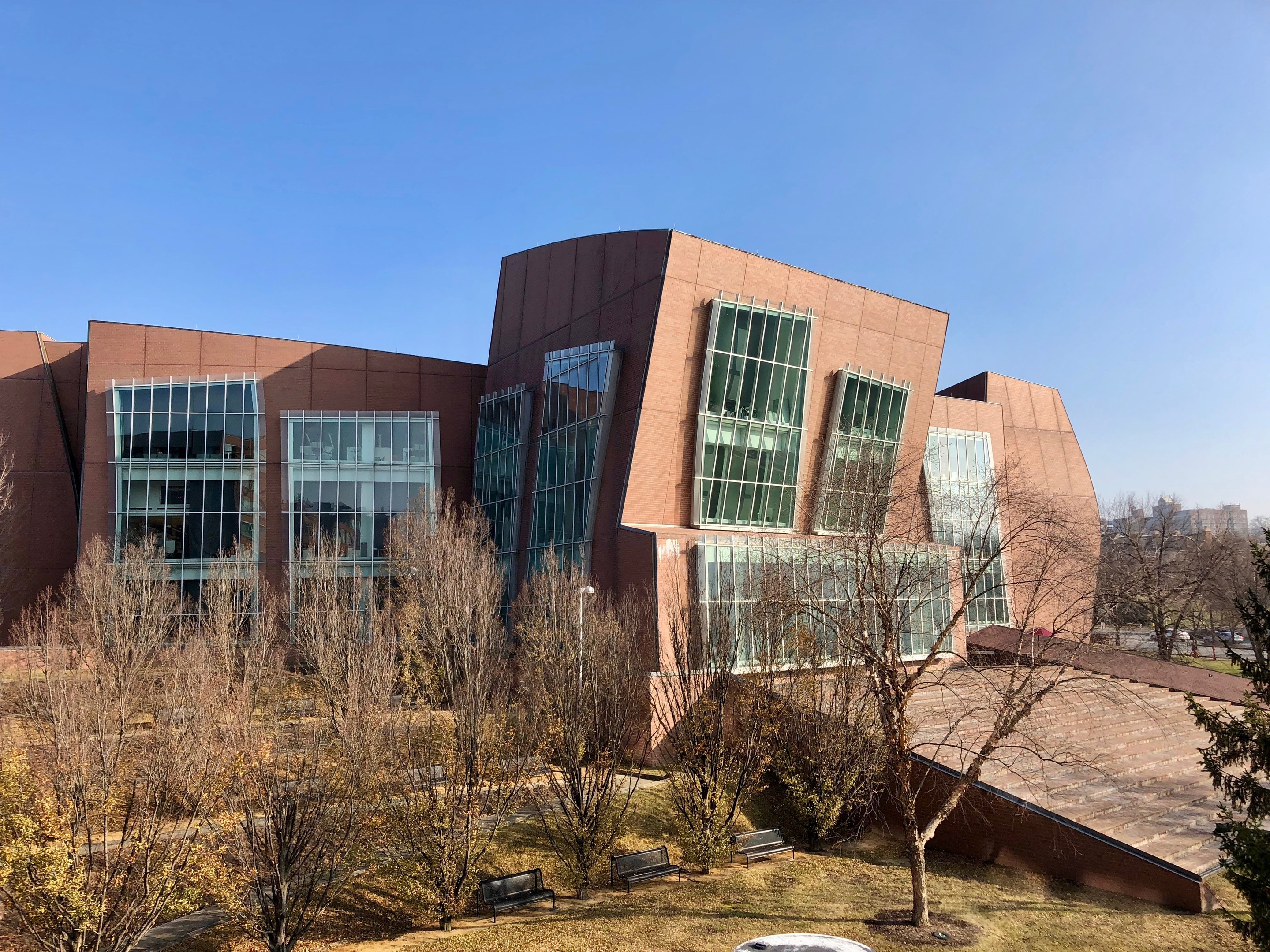 Großes modernes Gebäude mit vielen Fenstern, umgeben von Bäumen, Bänken, Gras und einem Laternenmast, vor einem klaren blauen Himmel mit anderen Gebäuden im Hintergrund, identifiziert als der neue Campus der University of Illinois at Chicago.