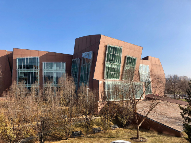 Großes modernes Gebäude mit vielen Fenstern, umgeben von Bäumen, Bänken, Gras und einem Laternenmast, vor einem klaren blauen Himmel mit anderen Gebäuden im Hintergrund, identifiziert als der neue Campus der University of Illinois at Chicago.