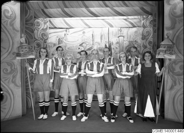 Schwarzes und weißes Foto einer Frauenfußballmannschaft der 1940er Jahre, die auf einer Bühne steht, lacht und Stöcke hält, mit einer bemalten Wand und Text am unteren Rand.