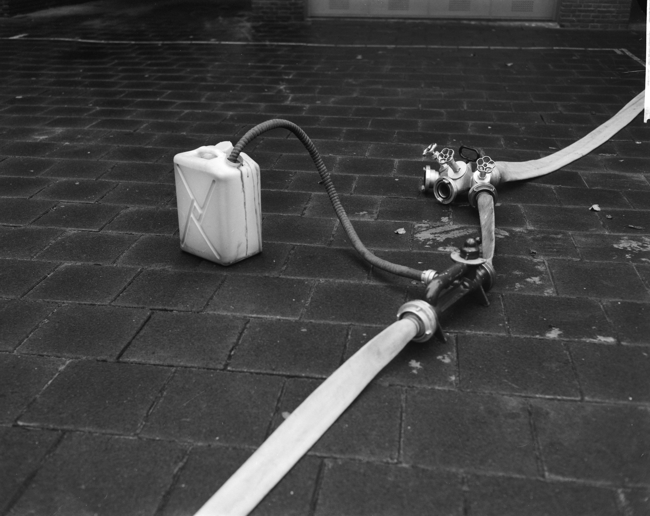 Schwarz-weißes Foto eines Gasbehälters mit einem Schlauch, der vor einem Gebäude mit einer Tür im Hintergrund auf dem Boden steht.
