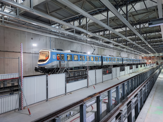 Blauer und weißer Zug fährt durch eine Indoor-Station mit einer Plattform auf der rechten Seite, einer Wand im Hintergrund und einer Decke mit Stangen und Lampen.