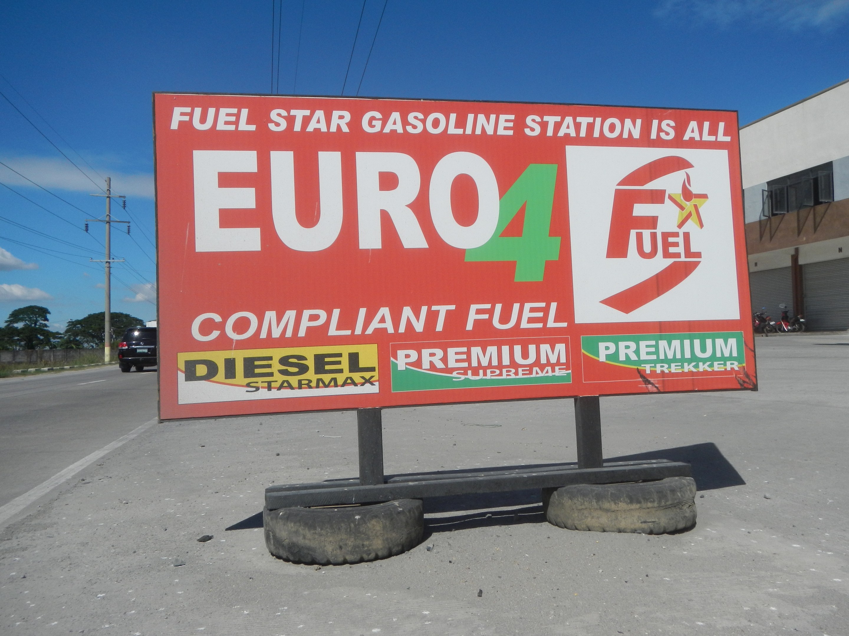 Eine Tankstelle mit einem "Euro4 Fuel"-Schild im Vordergrund, Fahrzeuge auf der Straße, Strommasten mit Drähten, Bäume, ein Gebäude auf der rechten Seite und ein sichtbarer Himmel.