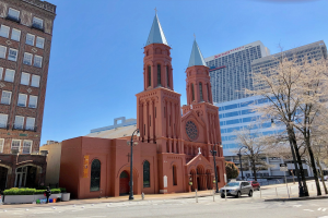 Die St. Patrick's Cathedral in Philadelphia, Pennsylvania, eine große rote Kirche mit einem Turm, steht in der Mitte einer Stadtstraße umgeben von Gebäuden, Straßenlaternen, Autos, Fußgängern, Büschen, Bäumen und unter einem klaren blauen Himmel.