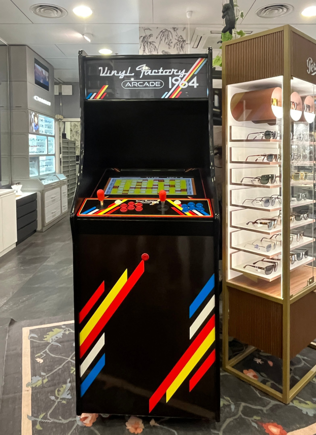 Ein vintage Arcade-Automat in der Mitte eines retro-stylischen Ladens mit einer Brillenhalterung auf der rechten Seite, Schränken, einem Fernseher und Deckenlampen im Hintergrund.