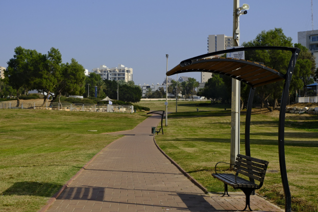 Eine Parkbank auf einer grünen Wiese umgeben von Bäumen, Masten, Lichtern und Gebäuden, mit einem Mülleimer im Vordergrund.