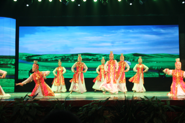 Eine Gruppe von Menschen in bunten Kostümen performs auf einer Bühne, beleuchtet von Scheinwerfern von oben und umgeben von Pflanzen, mit einem Bildschirm im Hintergrund.