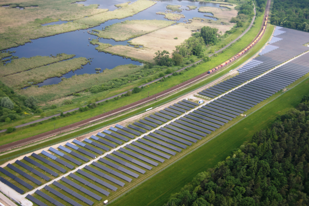 Eine Luftaufnahme eines Solarparks mit zahlreichen Solarpanelen in einem Feld, umgeben von Bäumen, Gras und Wasser, mit einem Zug, der auf einem nahen Bahngleis fährt.