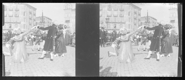 Schwarzes und weißes Foto einer Gruppe von Menschen in Kostümen, die auf der Straße tanzen, umgeben von Gebäuden, Straßenlaternen und Straßenlaternen unter einem klaren Himmel.
