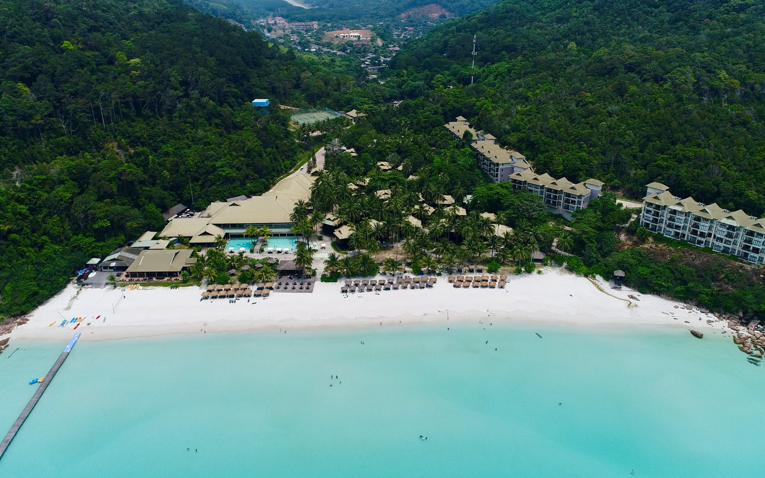Luftaufnahme eines Resorts am Strand in Koh Samui, Thailand, mit einer Brücke links, Gebäuden, Bäumen, Hügeln und einigen Menschen am Strand.