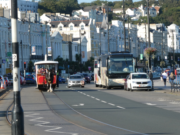 Eine belebte Stadtstraße mit hohen Gebäuden, Fahrzeugen, Fußgängern, Verkehrsampeln, Laternen, Schildern, Bäumen und einem klaren blauen Himmel.