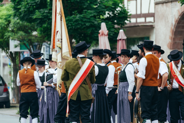 Gruppe von Menschen in traditioneller bayrischer Tracht, die eine Straße entlangmarschieren, einige halten Musikinstrumente und Fahnen, mit Bäumen, Gebäuden und einem Auto im Hintergrund.