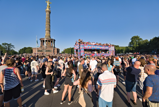 Große Menschenmenge versammelt sich vor einem Denkmal in Berlin mit Fahnen, einem säulenartigen Gebäude mit Statue und Inschrift, Bäumen und einem klaren blauen Himmel.