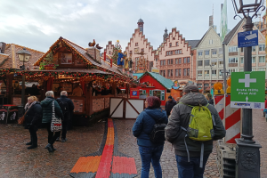Eine Gruppe von Menschen geht eine Kopfsteinpflasterstraße neben einem Weihnachtsmarkt in Nürnberg, Deutschland, entlang, mit Laternenmasten, Texttafeln und Gebäuden mit Fenstern im Hintergrund unter einem bewölkten Himmel.