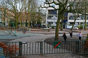 Eine Gruppe von Kindern spielt in einem Park mit einem Metallzaun, einer Bank, Pflanzen, einem Schild, Pfosten, einem Metallrahmen, Fahrzeugen, Menschen auf einem Weg, Bäumen, Gebäuden mit Fenstern und einem bewölkten Himmel.