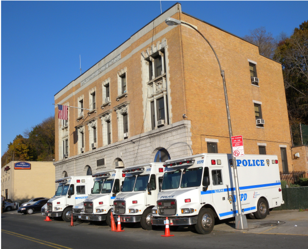 Mehrere Polizei-Lkw sind vor einem Gebäude mit Fenstern, einem Lichtmast, Absperrbaken, einem Schild, einem Zaun, Bäumen und einer Fahne mit einem Mast geparkt. Unten im Bild steht Text.