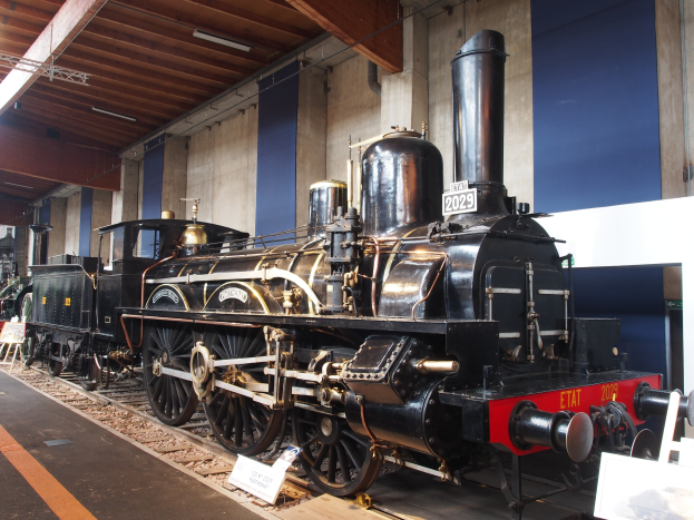 Alte Dampflokomotive in einem Museums-Setting mit umliegenden Eisenbahnschienen, Informationsschildern und anderen Ausstellungen, vor einer Wand mit Deckenbeleuchtung.