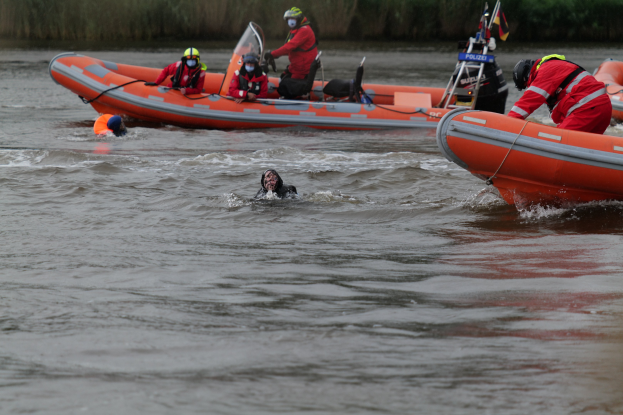 Menschen in Schwimmwesten und Helmen in einem aufblasbaren Boot auf einem Fluss, mit zwei Personen im Wasser im Vordergrund und Vegetation im Hintergrund.