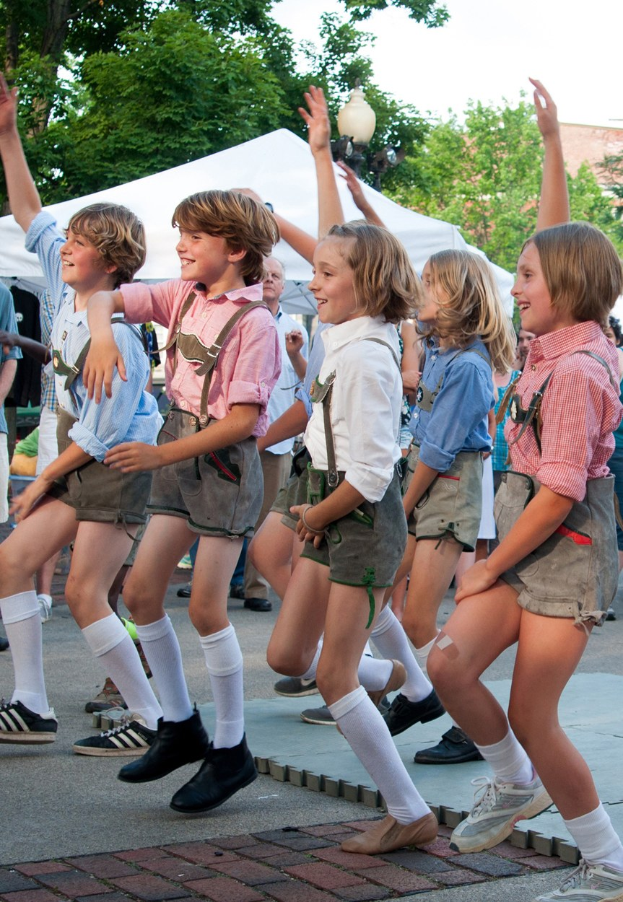 Kinder in traditioneller bayrischer Tracht, die auf einer Straße tanzen, umgeben von einer Menge, Zelten, Laternenmasten, Bäumen, Gebäuden und einem klaren blauen Himmel. Einige Kinder tragen Shorts und Schuhe.