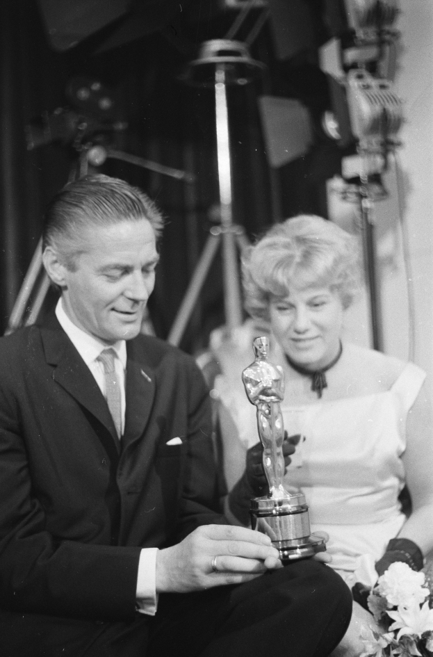 Ein Mann in einem schwarzen Blazer, einem weißen Hemd und einer Krawatte h├Ąlt eine Oscar-Statue in der Hand, waehrend er neben einer Frau in Abendgarderobe sitzt, die einen Blumenstrau├č h├Ąlt, mit weicher Beleuchtung im Hintergrund.