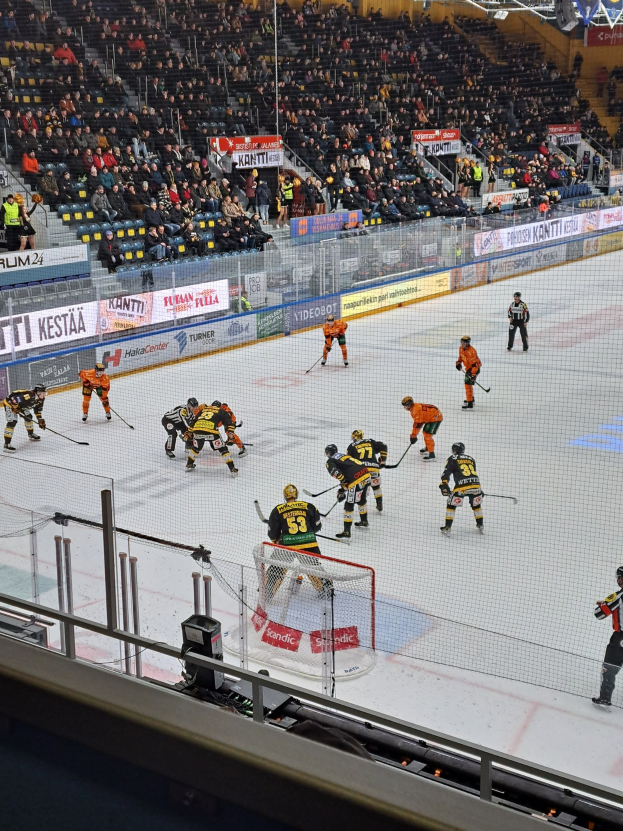 Gruppe von Menschen, die Hockey auf einem Eisring spielen, umgeben von einem Zaun, mit einem Netz in der Mitte, Zuschauern im Hintergrund, Schildern und Überdachungsbeleuchtung.