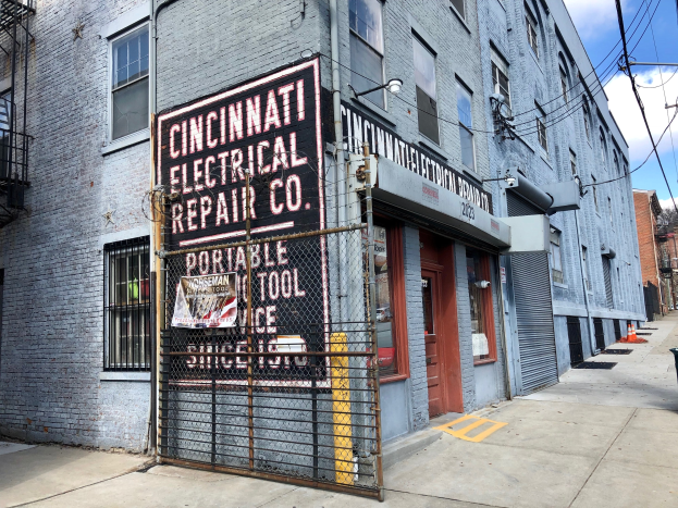 Außenansicht des Cincinnati Electrical Repair Co. Gebäudes mit einer Hinweistafel, einem Metallzaun, einem Müllcontainer, Pfählen, Drähten, Bäumen und einem bewölkten Himmel.