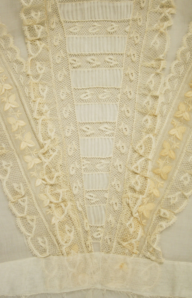 Nahaufnahme eines weißen Kleides mit zarter, kunstvoller Spitzenverzierung in verschiedenen Formen und Mustern auf einem leichten, luftigen Stoff, geeignet für den Sommer.