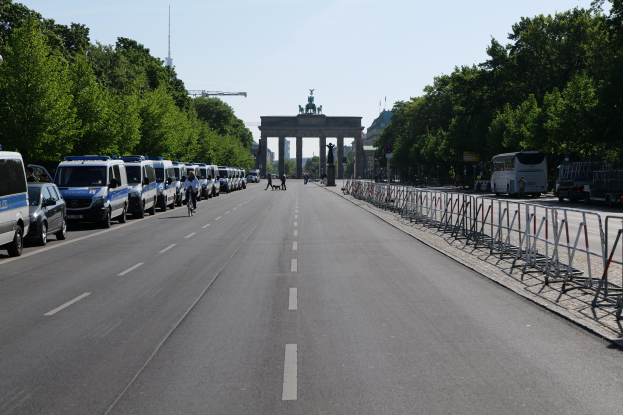 Eine lange Reihe von Polizeiwagen, die auf der Seite einer Straße vor dem Brandenburger Tor in Berlin, Deutschland, geparkt sind, mit Menschen, die Fahrräder fahren und auf der Straße stehen, Absperrungen und Bäume, die die Seiten säumen, und ein Tor mit Statuen im Hintergrund.