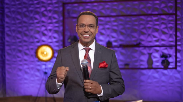 Stephen Colbert, Moderator von 'The Daily Show with Trevor Noah', steht auf einer Buhne und halt einen Mikrofon, tragt einen Anzug, Krawatte und Brille, mit einem Licht und Stander hinter ihm.