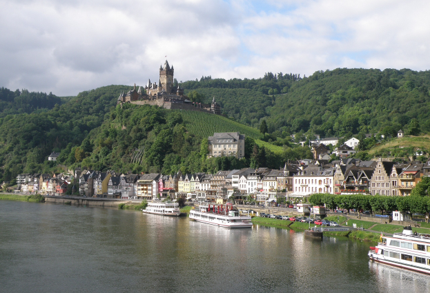 Ein idyllischer Blick auf den Rhein in Deutschland mit einer Burg auf einem Hügel, Booten auf dem Fluss, Fahrzeugen auf einer näheren Straße und einem bewölktem Himmel.