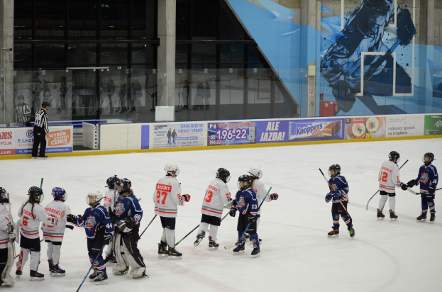 Junge Menschen in Helmen und Hockey-Ausrüstung spielen Eis-hockey auf einer Indoor-Eisfläche mit Bannern, Säulen und Glaswänden im Hintergrund.