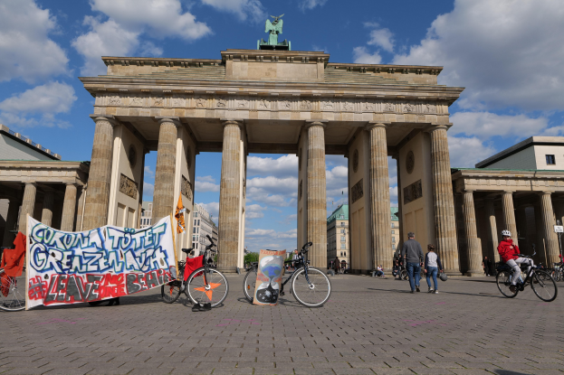 Gruppe von Menschen, die Fahrräder vor dem Brandenburger Tor in Berlin fahren, mit dem Torbogen, Säulen und Statue im Vordergrund, einem Banner mit Text, Gebäuden, Bäumen und einem bewölkten Himmel im Hintergrund.