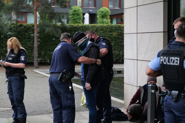 Eine Gruppe von Polizeibeamten steht vor einem Gebäude mit einer Frau, die ein Buch und einen Stift hält, während andere in der Nähe sitzen oder stehen, mit Bäumen und Gebäuden im Hintergrund.