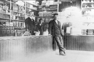 Drei Männer stehen an einem Tresen in einem Laden mit verschiedenen Gegenständen, während ein Regal mit Büchern und anderen Gegenständen im Hintergrund zu sehen ist, während eines Überfalls.