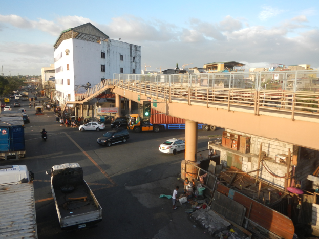 Eine belebte Stadtstra├če mit Autos, Lastwagen, Fü├čgängern, einer Brücke mit Gel├ädern und Pfeilern, Ampeln, Geb├Ąude mit Fenstern, B├Ąumen und einem bew├Âlktem Himmel.