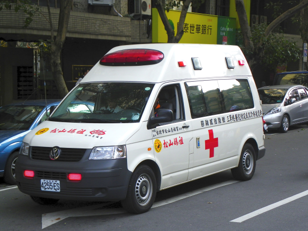 Ambulanz auf der Seite der Straße mit anderen Fahrzeugen, Bäumen, Gebäuden, Texttafeln und Klimaanlagen im Hintergrund.