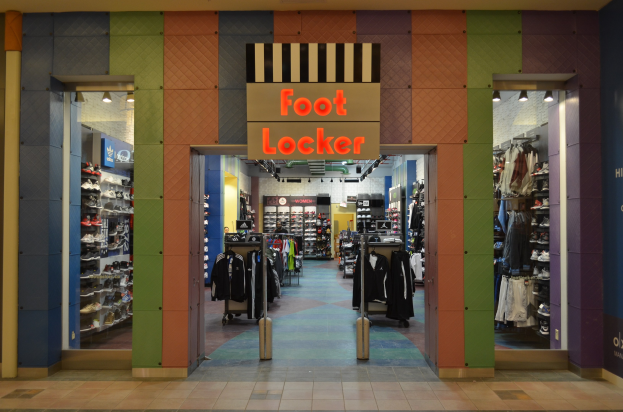Innenansicht eines Foot Locker-Schuhgeschäfts mit Kleidung an Bügeln und Ständern unter Geschäftbeleuchtung.