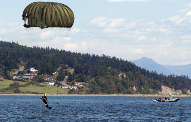 Eine Person beim Drachenfliegen über einem Gewässer mit einem Boot auf der rechten Seite, Bäumen, Gebäuden, Hügeln und einem klaren blauen Himmel im Hintergrund.