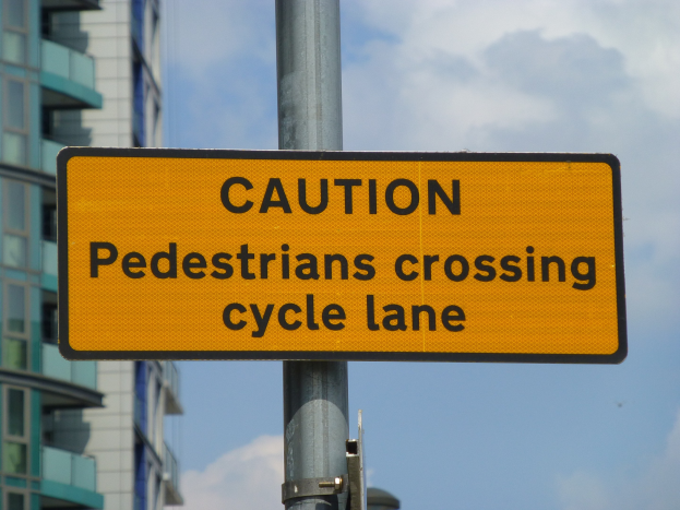 Gelbes Warnschild an einem Pfahl vor einem Gebäude mit einem klaren blauen Himmel im Hintergrund, das "Fußgänger überqueren Radweg" anzeigt.